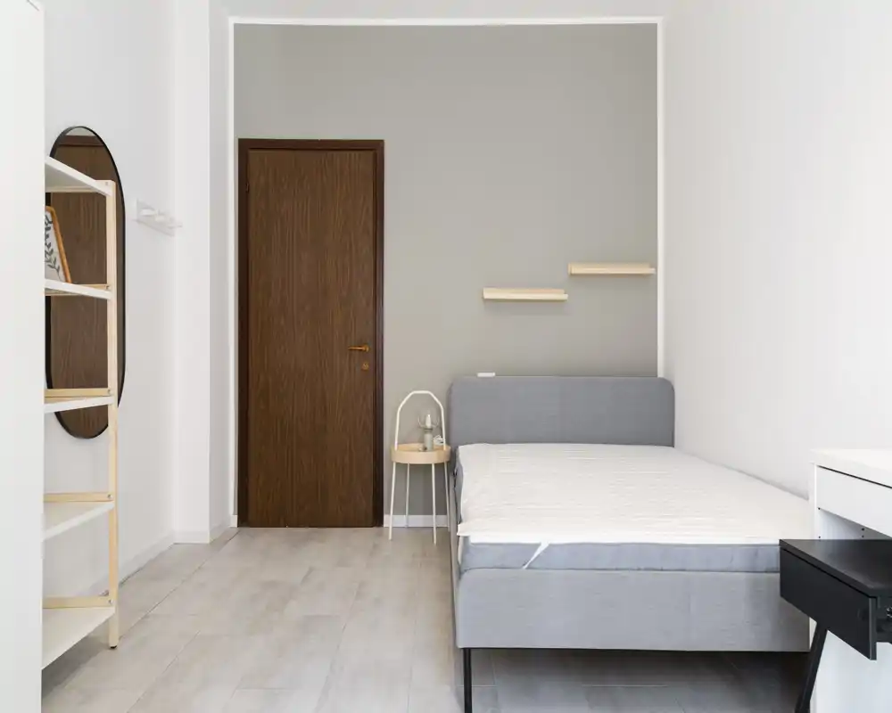 Minimalist, bright bedroom with a full gray bed, small round bedside table and simple wall shelves — ideal for a clean, modern rental listing.