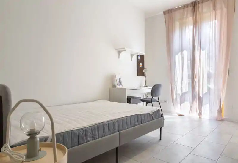 Bright, minimalist bedroom with a comfortable double bed, a small desk and soft pink curtains letting in natural light — ideal for a peaceful stay.