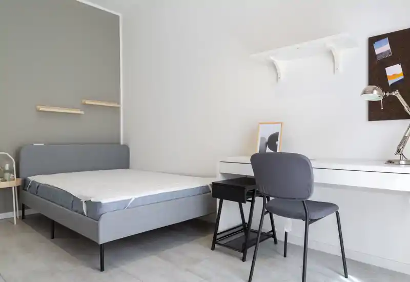 Bright, minimalist bedroom with a gray upholstered bed and a white study desk. The room features clean lines, neutral tones and good natural light, ideal for a single occupant or student.