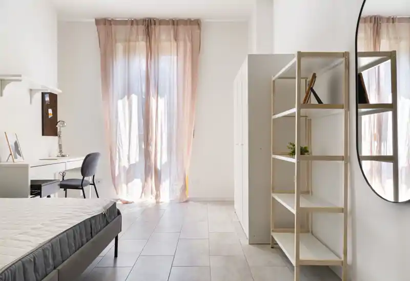 Bright, modern bedroom with a neatly made bed, a study corner with desk and chair, open shelving and soft sheer curtains admitting natural light.