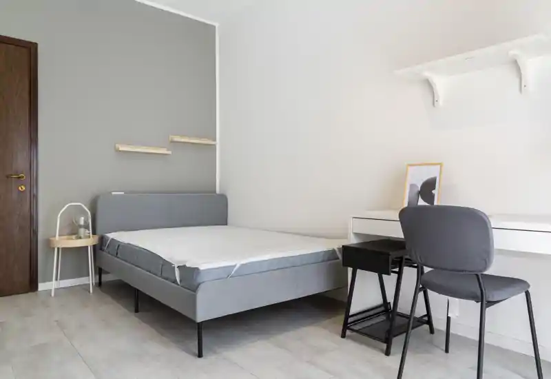 Bright, minimal bedroom with a grey upholstered double bed, bedside table and a small desk with chair — modern, clean styling and soft natural light.