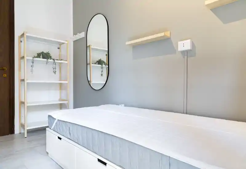 Bright, minimal bedroom with a neatly made single bed, wall-mounted shelves and an oval mirror that enhance the sense of space.