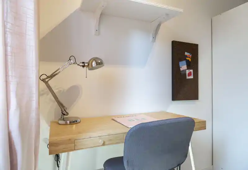 Angolo della camera utilizzato come studio: scrivania in legno con lampada da tavolo, bacheca e sedia imbottita, luce naturale morbida da sinistra.