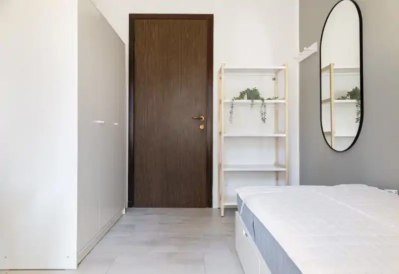 Bright, minimalist bedroom view showing a neatly made bed, wardrobe, shelving with plants and an oval wall mirror — clean, modern styling ideal for rental photos.
