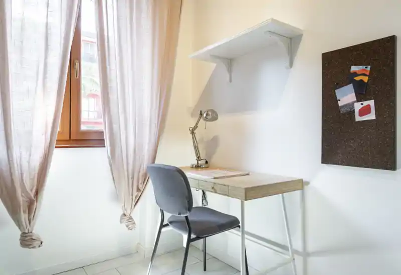 Small, bright workspace corner with a wooden desk, task lamp and upholstered chair next to a curtained window — a neat study or home office nook.