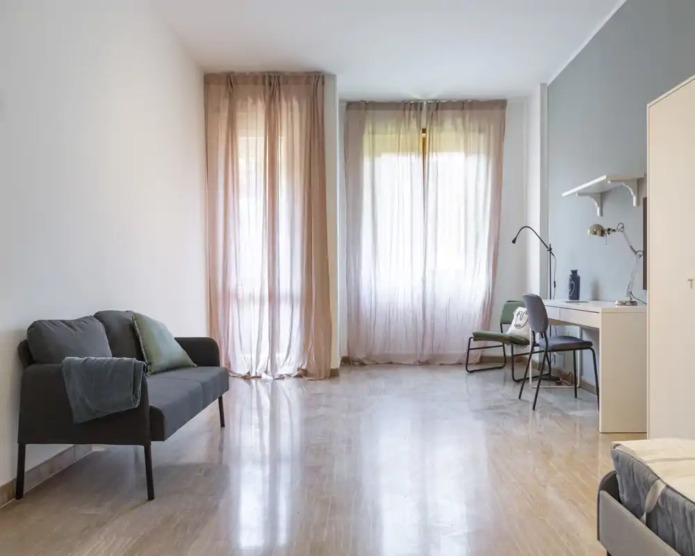 Spacious, bright living area with a small sofa, desk and sheer curtains providing soft natural light. Minimal, modern styling makes the room feel airy and functional for both relaxing and working.