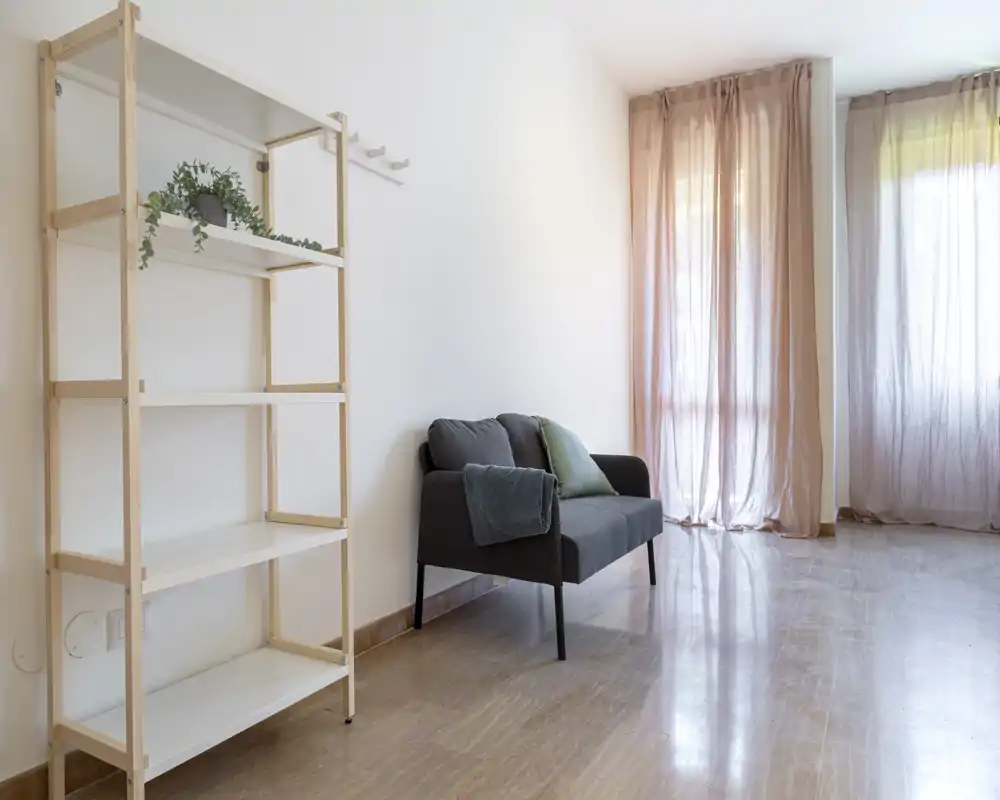Minimal, well-lit living area with a compact two-seat sofa, light shelving and soft curtains creating a calm, airy atmosphere.