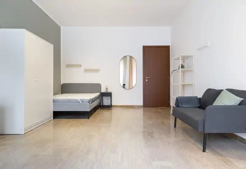 Dormitorio luminoso y minimalista con cama doble, armario y una pequeña zona de estar con sofá. Líneas limpias, tonos neutros y amplio suelo libre, perfecto para fotografías de alquiler.