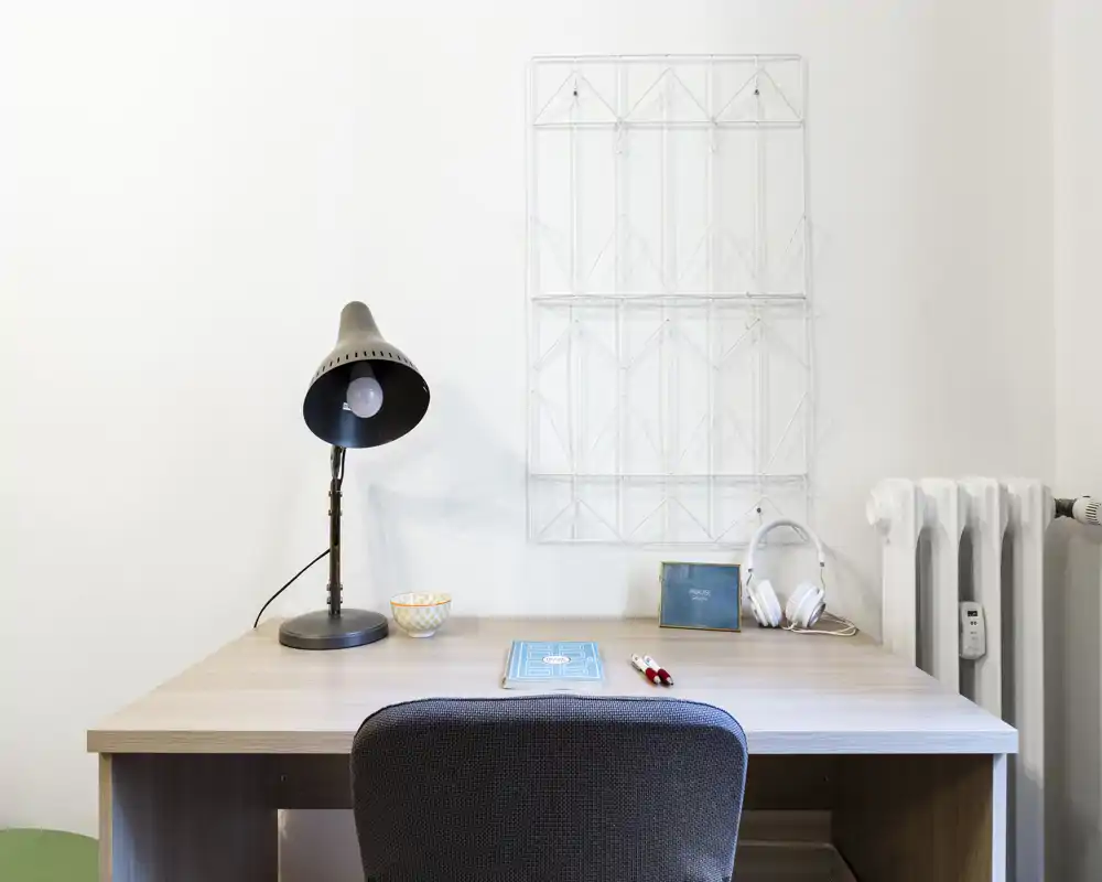 Angolo studio ordinato in camera da letto con scrivania in legno, lampada, sedia e piccoli accessori su una parete neutra — perfetto come postazione di studio.