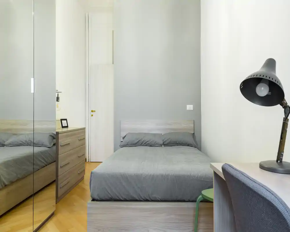 Camera da letto moderna e luminosa con letto matrimoniale centrale, biancheria grigio neutro, armadio con specchi e una piccola scrivania con lampada — pulita e pronta per gli ospiti.