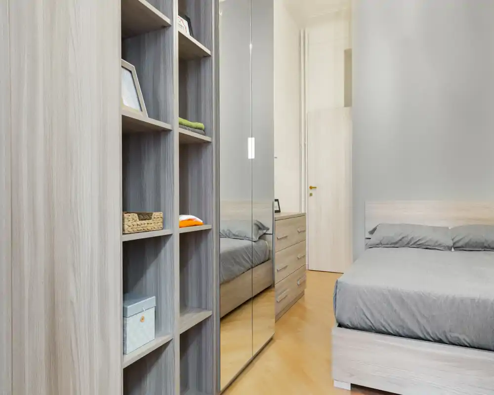 Camera da letto moderna e luminosa con letto matrimoniale ben rifatto, armadio con ante a specchio e scaffalatura a vista—stile minimal e funzionale per affitti.