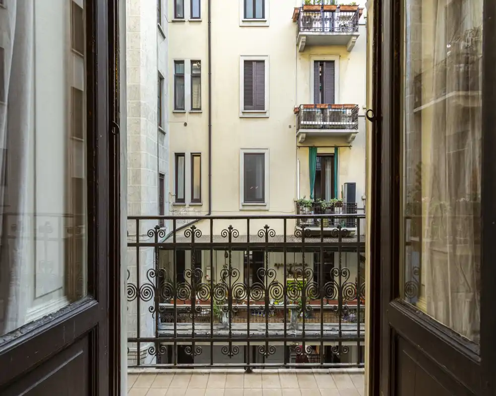 Piccolo balcone privato che si affaccia su un cortile interno con ringhiera in ferro battuto e facciate degli edifici vicini; pavimento piastrellato e porte a doppia anta incorniciano la vista.