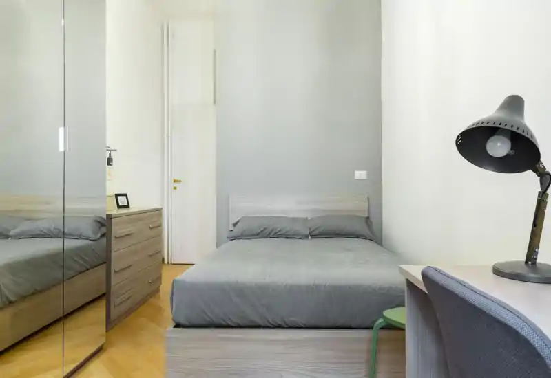 Camera da letto moderna e luminosa con letto matrimoniale centrale, biancheria grigio neutro, armadio con specchi e una piccola scrivania con lampada — pulita e pronta per gli ospiti.