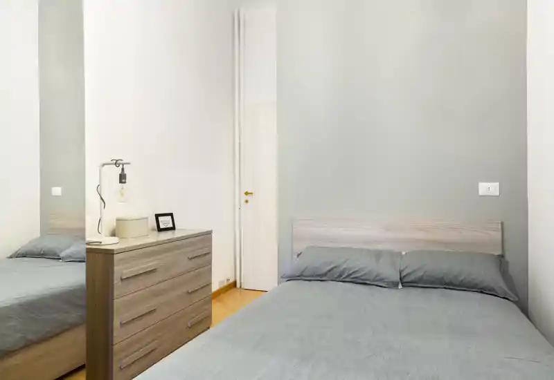 Camera da letto ordinata e luminosa con letto matrimoniale, testiera in legno chiaro e comò abbinato; biancheria grigia e illuminazione soffusa creano un'atmosfera moderna e rilassante, perfetta per annunci.