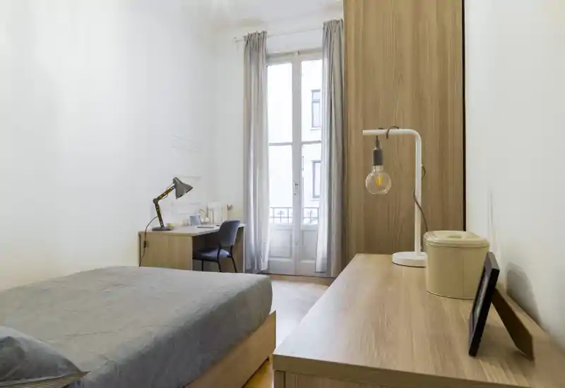 Camera da letto luminosa e minimal con letto matrimoniale, mobili in legno e una scrivania vicino alla finestra alta che lascia entrare luce naturale. Linee pulite e tessuti morbidi creano uno spazio funzionale e rilassante.