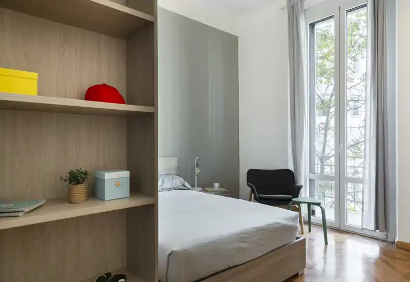 Camera da letto luminosa e minimale con ampia finestra, letto ordinato e una poltrona accogliente — perfetta per un soggiorno tranquillo in città.