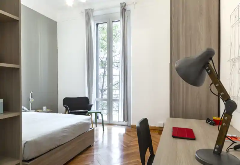 Camera da letto luminosa e moderna con letto matrimoniale, scrivania e una finestra alta che lascia entrare molta luce naturale. Pavimenti in parquet, una poltrona confortevole e un'area studio semplice — perfetta per studenti o professionisti.