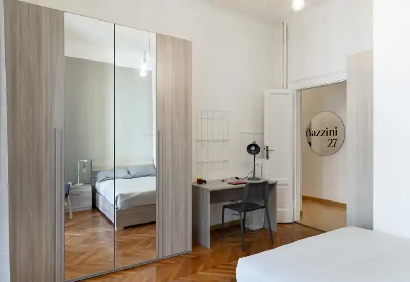 Camera da letto moderna e luminosa con letto matrimoniale, armadio con ante a specchio e una piccola scrivania; la luce naturale e il parquet caldo creano un’atmosfera accogliente.