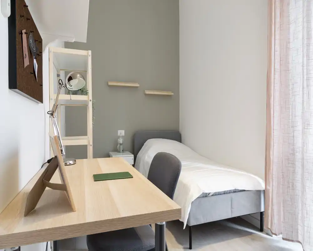 Camera singola compatta e ben arredata con letto ordinato, comodino e scrivania in legno vicino alla finestra che illumina naturalmente — ideale per uno studente o una persona sola.