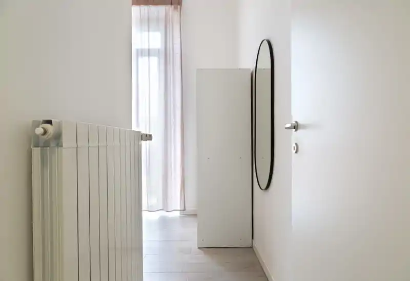 Minimal, well-lit entrance hallway with a slim radiator, a tall narrow cabinet and an oval wall mirror reflecting light from a curtained window.