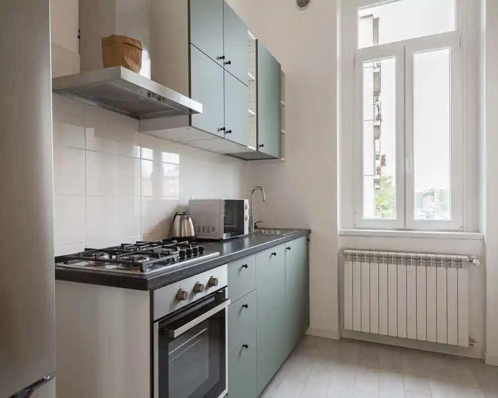 Cucina moderna e luminosa con pensili verde menta, piano cottura a gas, forno e ampia finestra che offre luce naturale. Piano di lavoro pulito con microonde e bollitore, perfetta per appartamenti compatti.