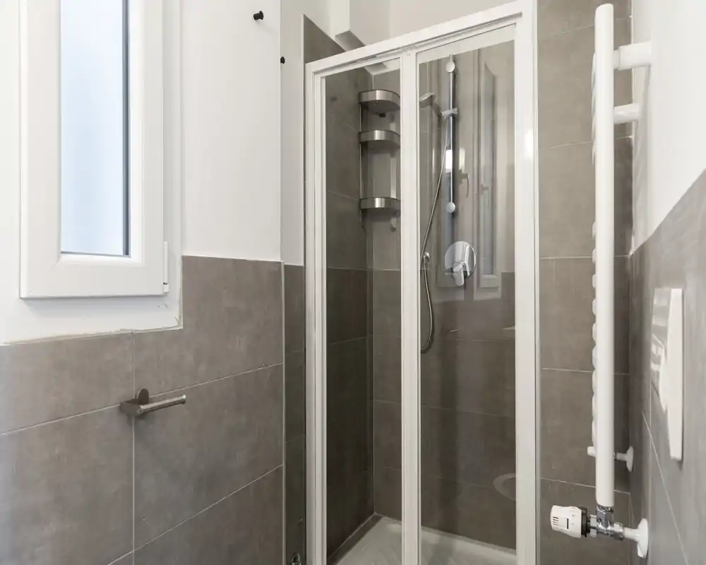 Bagno moderno compatto con box doccia completamente chiuso, piastrelle grigie e termoarredo; ambiente luminoso, pulito e dal design minimale.