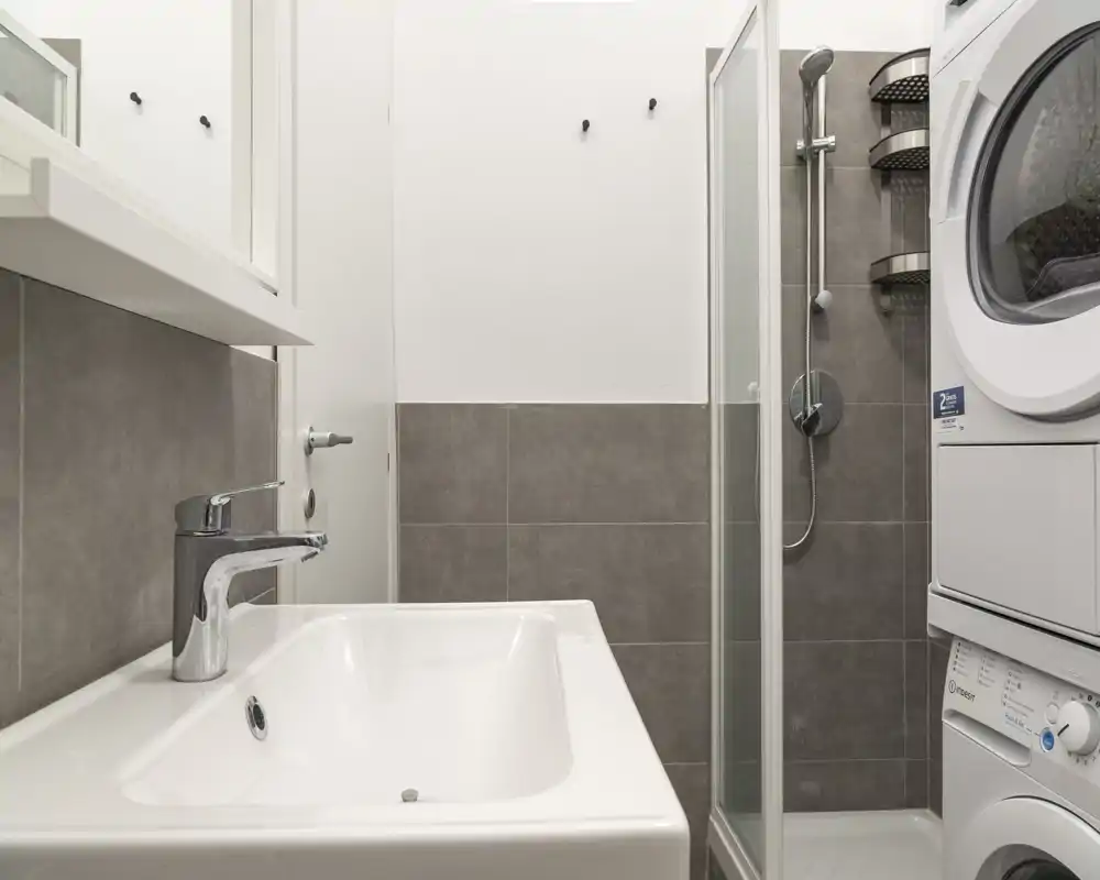 Bagno moderno e compatto con lavabo in primo piano, doccia con box in vetro e colonna lavatrice-asciugatrice. Piastrelle grigie neutre e sanitari bianchi donano un aspetto pulito e funzionale.