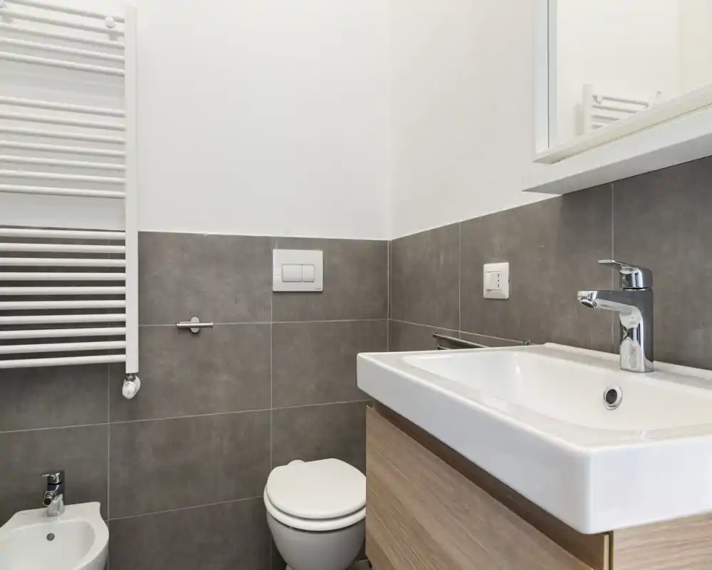 Bagno moderno e compatto con lavabo a muro, rubinetteria cromata, WC e bidet; piastrelle grigie a mezza altezza e scaldasalviette. Design pulito, luminoso e minimal ideale per un annuncio.