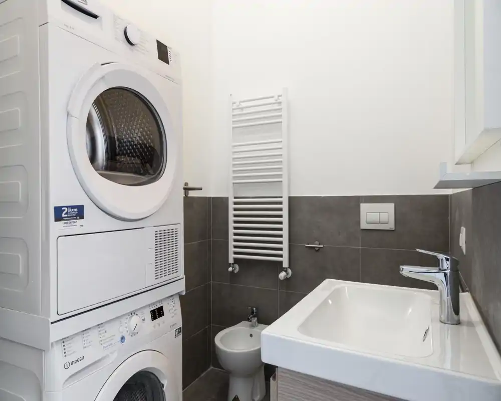 Bagno moderno compatto con lavatrice e asciugatrice sovrapposte, termoarredo a parete, bidet e lavabo con rubinetto cromato; piastrelle nei toni neutri e pareti bianche per un ambiente pulito e funzionale.