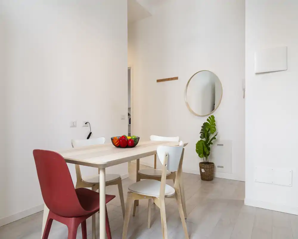 Zona pranzo luminosa e minimal con tavolo in legno chiaro, sedie miste (con una sedia rossa come accento) e una ciotola di frutta decorativa; uno specchio e una pianta in vaso donano calore.