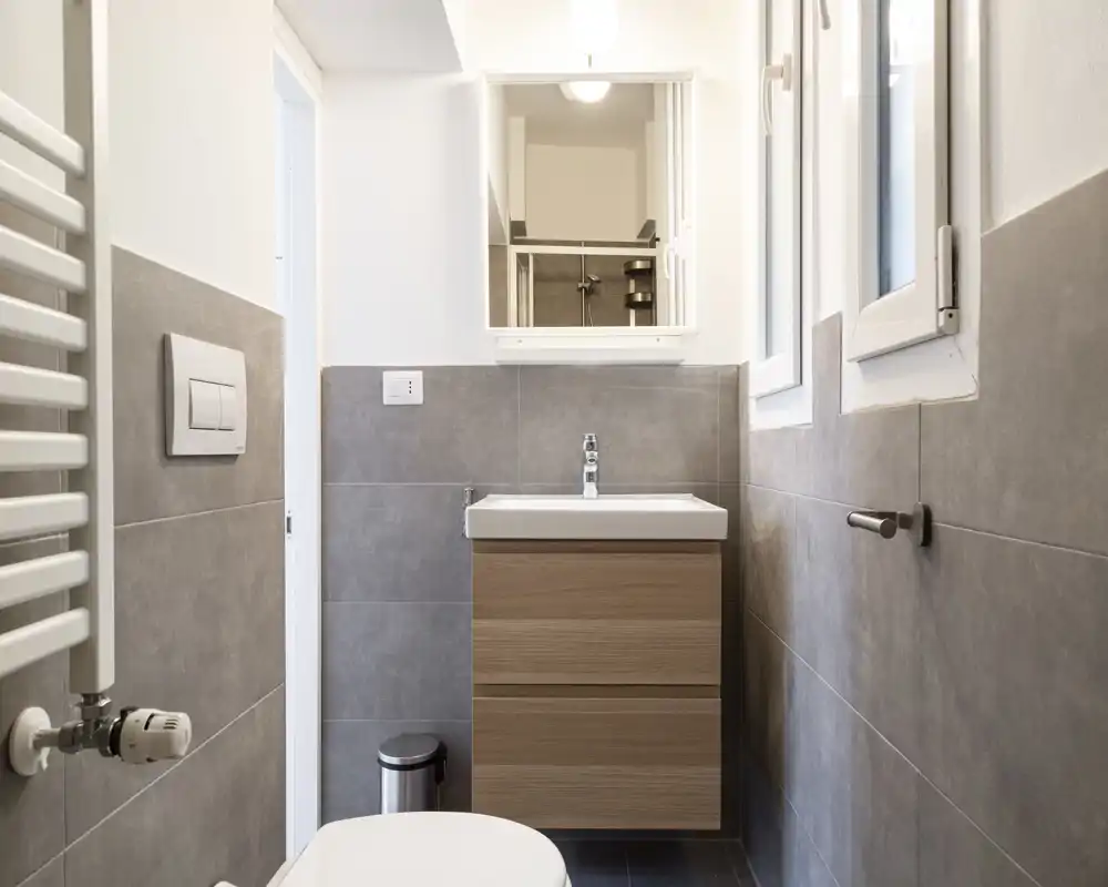 Bagno moderno e compatto con piastrelle grigie, mobile lavabo in legno a parete, specchiera, wc e termosifone porta-asciugamani — pulito e ben illuminato.