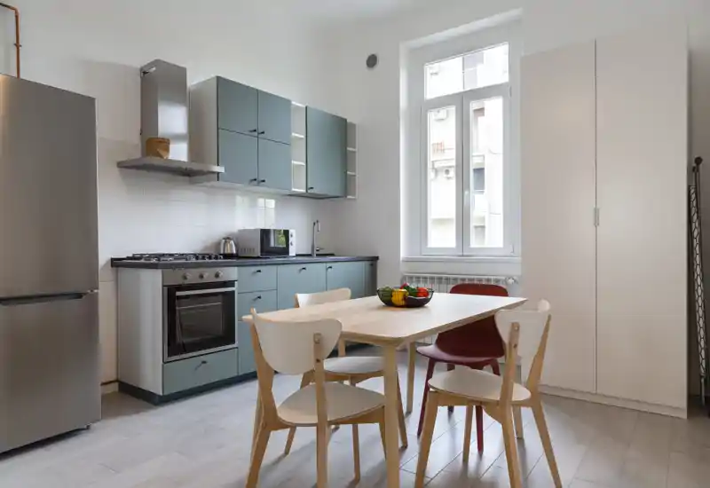 Cocina moderna y luminosa con muebles inferiores verde azulado, electrodomésticos de acero inoxidable y mesa de madera para cuatro—iluminada por una gran ventana y decorada con un cuenco de frutas.