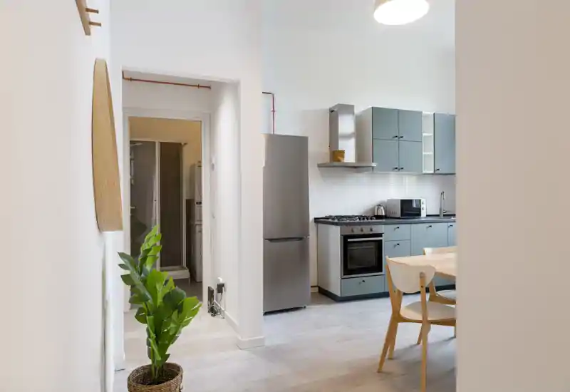 Cocina compacta y moderna con muebles verde azulado, electrodomésticos de acero inoxidable y un pequeño comedor; luminosa, ordenada y decorada con una planta que aporta calidez.