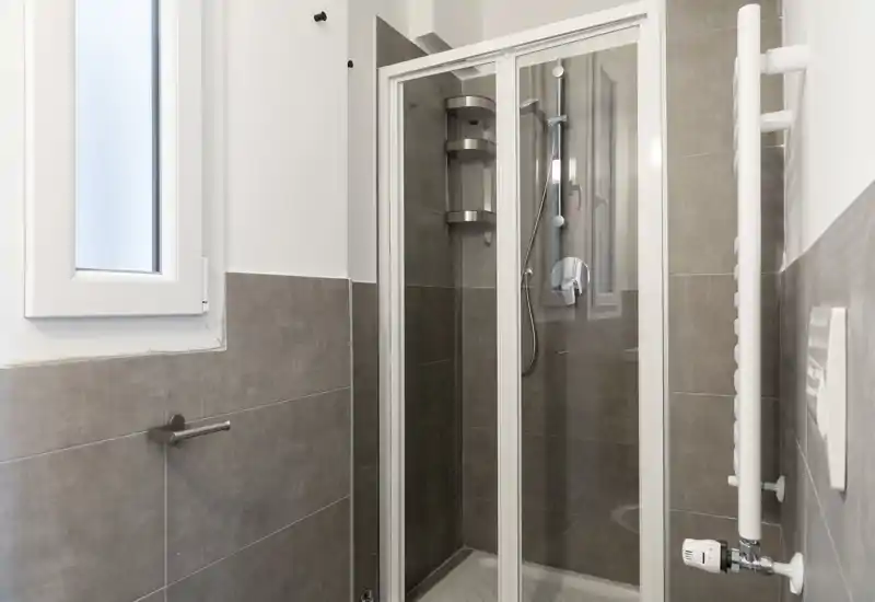 Baño moderno y compacto con ducha acristalada, azulejos grises, pequeña ventana translúcida y radiador toallero vertical.