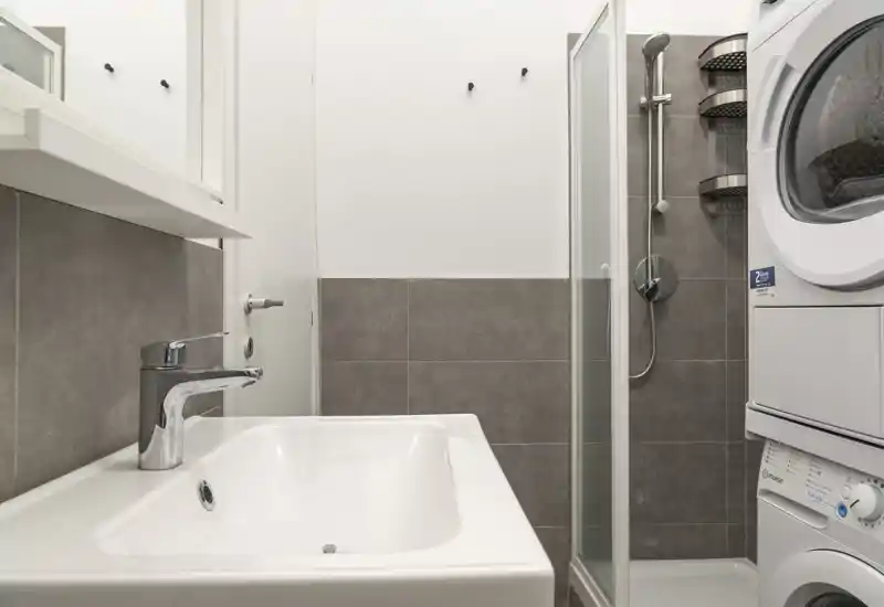 Baño moderno y compacto con lavabo blanco, mampara de ducha de cristal y lavadora-secadora apiladas; azulejos grises y grifería limpia que ofrecen un espacio funcional y bien iluminado.