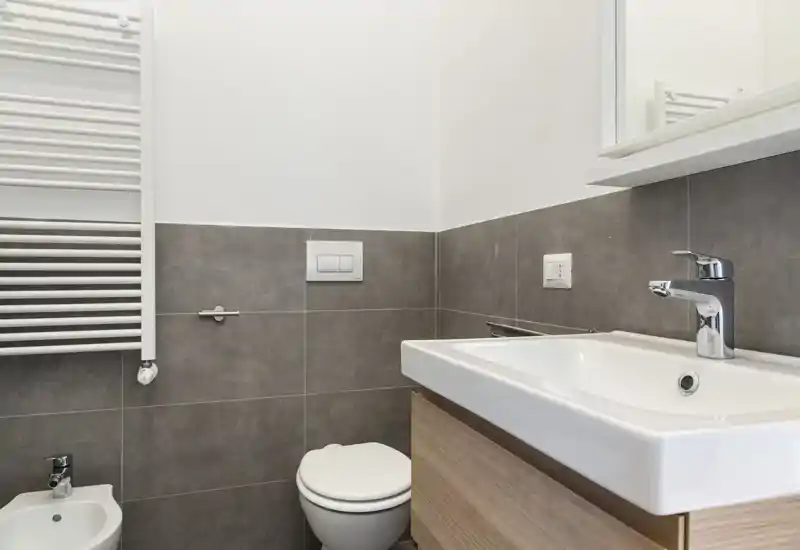 Baño moderno y compacto con lavabo mural, grifo cromado, inodoro y bidé; azulejos grises a media altura y toallero calentador. Diseño limpio, luminoso y minimalista, ideal para un anuncio de alquiler.