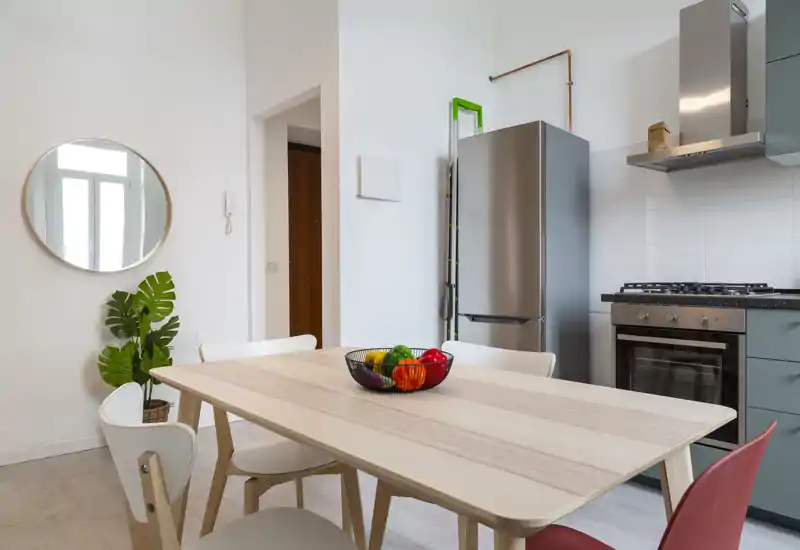 Cocina-comedor moderna y luminosa con mesa de madera clara, sillas mezcladas y un cuenco de frutas; frigorífico y horno de acero visibles, creando un ambiente de comedor abierto y acogedor.
