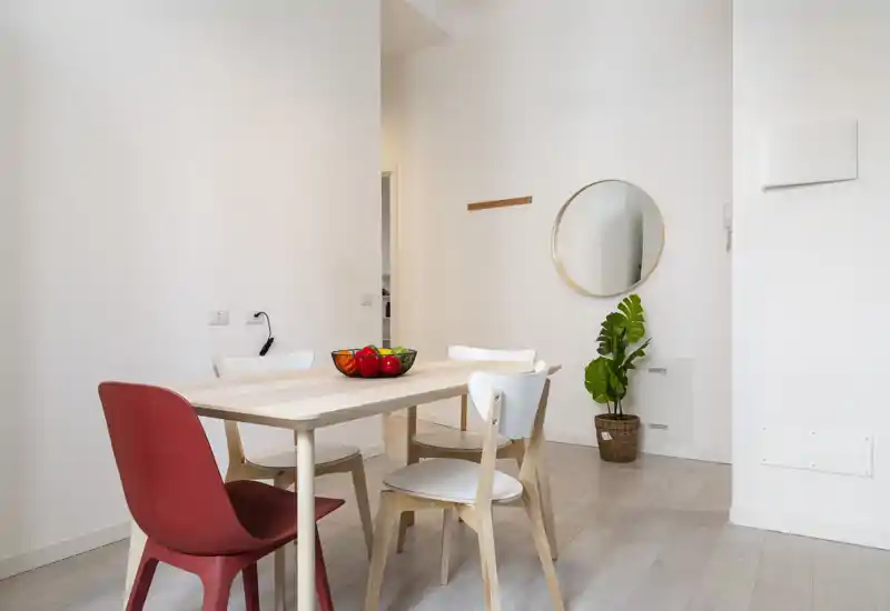 Zona de comedor minimalista con mesa de madera clara, cuatro sillas (incluida una silla roja de acento), un cuenco de frutas decorativo y una planta en maceta, sobre paredes blancas y un espejo redondo.
