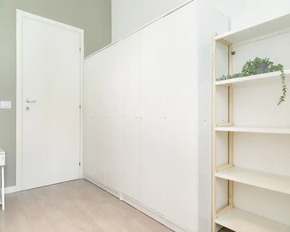 Ingresso/corridoio minimale con armadi a muro bianchi e mensole aperte. Pavimento chiaro e porta bianca chiusa creano uno spazio di ingresso ordinato.