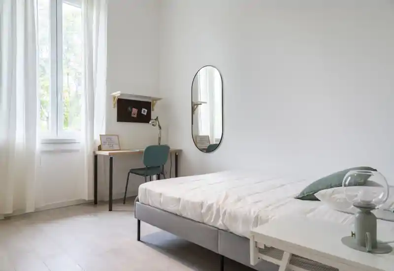 Chambre lumineuse et minimaliste avec un lit bien fait, table de chevet avec lampe et un petit bureau près de la fenêtre apportant la lumière naturelle — un espace de sommeil calme et aérien.