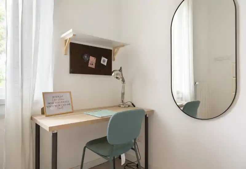 Petit espace de travail lumineux près d'une fenêtre avec un bureau en bois simple, une chaise bleu-vert, une lampe de bureau et un grand miroir ovale reflétant une partie de la pièce.
