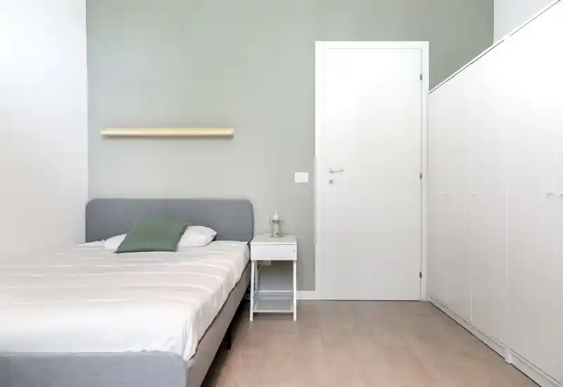 Chambre minimaliste avec un lit gris rembourré, table de chevet blanche et placards encastrés blancs. Mur vert pâle et coussin apportent une atmosphère calme et moderne.