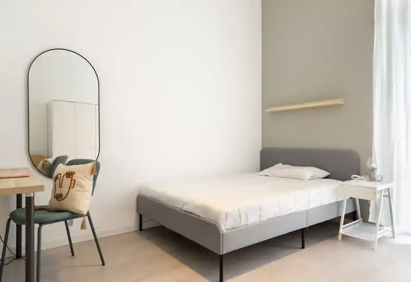 Bright, minimal bedroom with a double bed, bedside table and a desk area; neutral tones and soft natural light create a calm atmosphere.