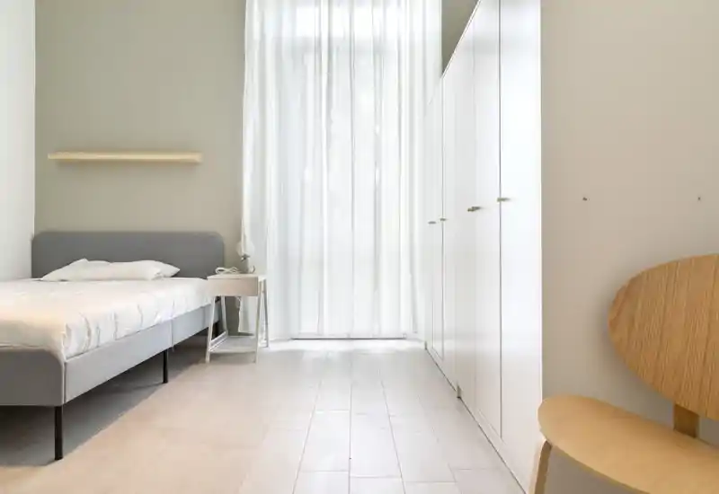 Minimal, bright bedroom with a single bed, bedside table and large wardrobe by a sheer-curtained window; soft natural light and pale wood floors create a calm, airy atmosphere.
