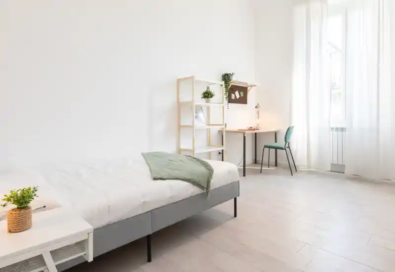 Bright, minimalist bedroom with a neatly made single bed, a small bedside table with plant, and a simple desk by a large window letting in abundant natural light.