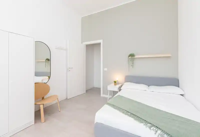 Bright, minimalist bedroom with a double bed dressed in white linens and a soft green throw, bedside table with a lamp and a small plant, wardrobe and a wall mirror create a calm, contemporary space.