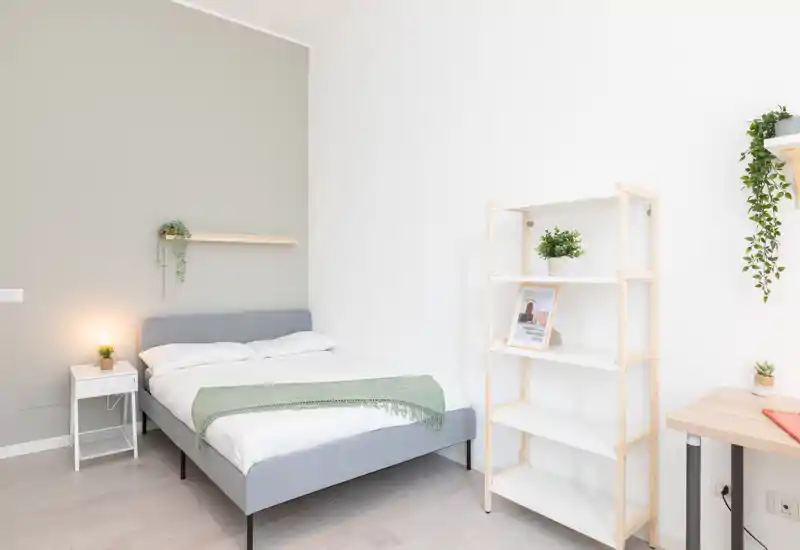 Bright, minimal bedroom with a neatly made double bed, bedside table and shelving; modern neutral tones and small plants create a calm, welcoming atmosphere.