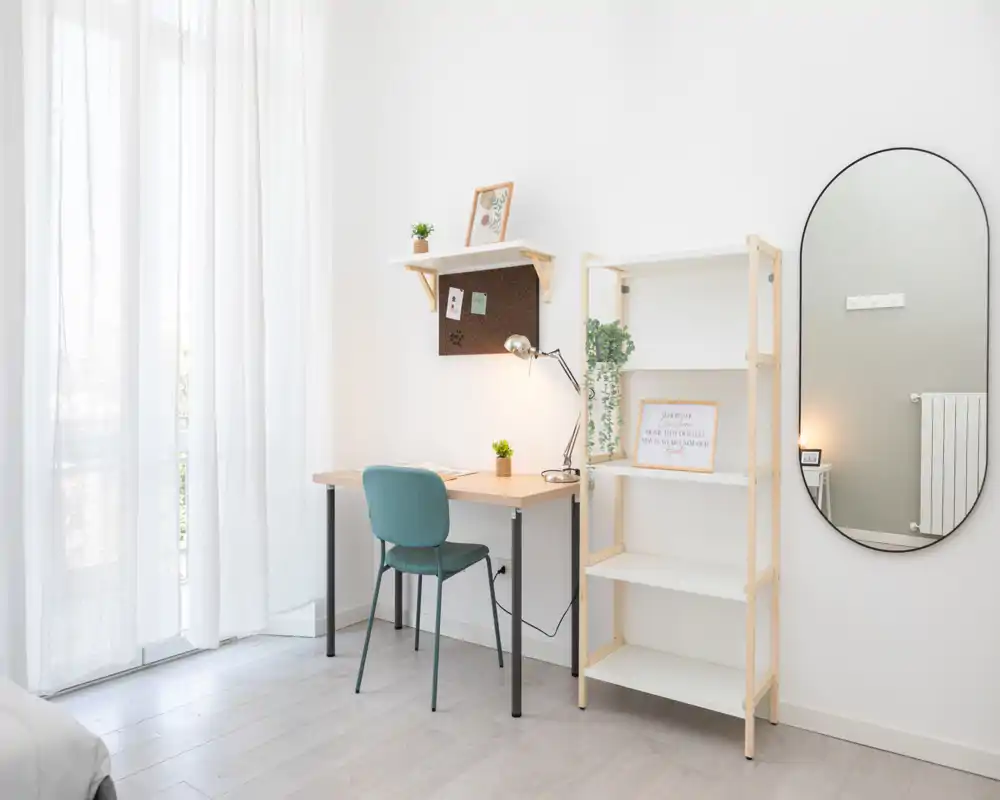 Bright, minimalist bedroom corner with a study desk, shelving and a tall oval mirror; sheer curtains provide soft natural light and the space feels calm and uncluttered.