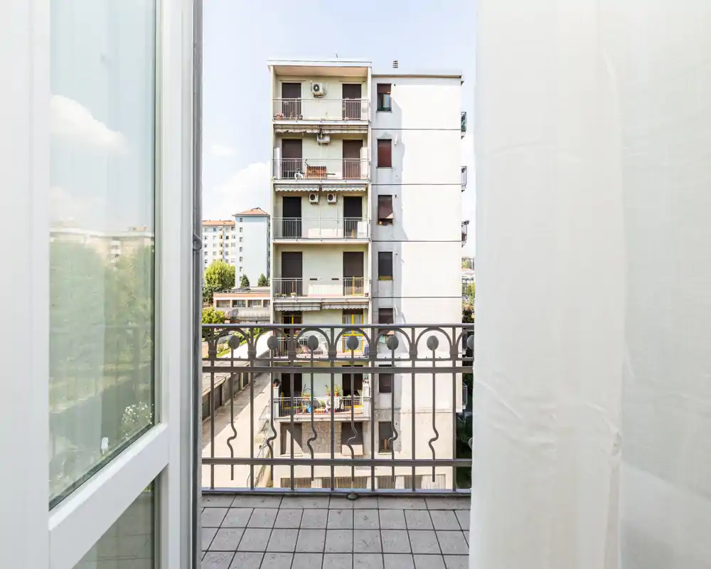Small private balcony view looking towards neighboring apartment blocks; tiled floor, decorative metal railing and light incoming through open doorway.