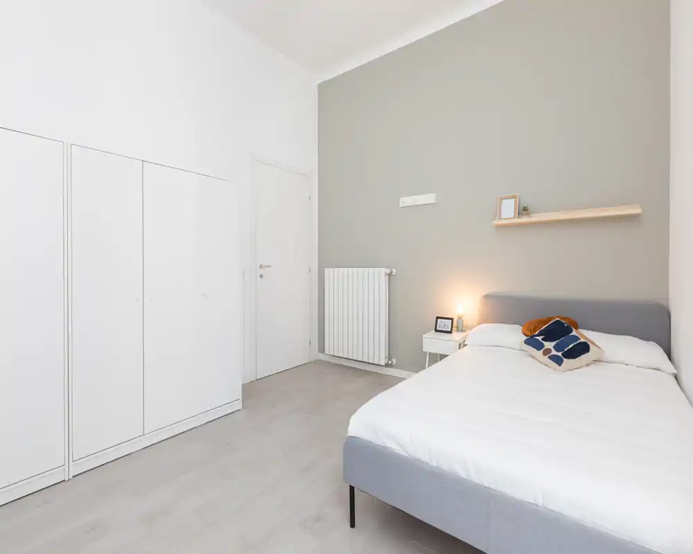 Bright, minimal bedroom with a comfortable double bed, bedside table with lamp and a simple wall shelf — ideal for a modern rental.